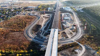 O vedere aeriană prezintă o intersecție de autostradă în construcție cu drumuri de asfalt, zone de pământ și utilaje de șantier.