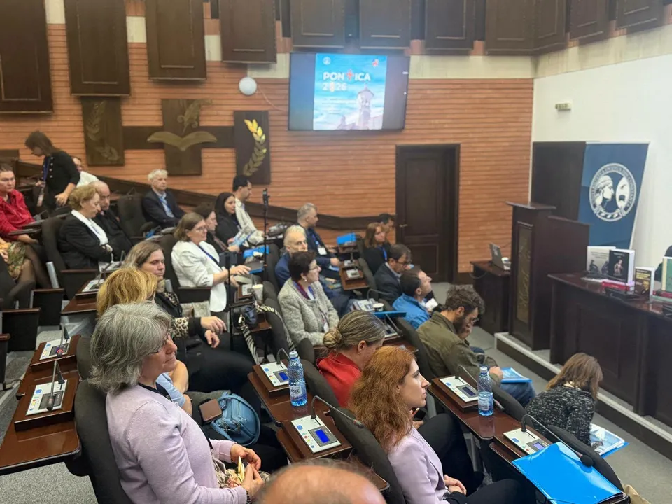 Oamenii stau așezați în rânduri de scaune într-o sală de conferințe, privind spre un ecran de proiecție suspendat pe un perete de lemn.