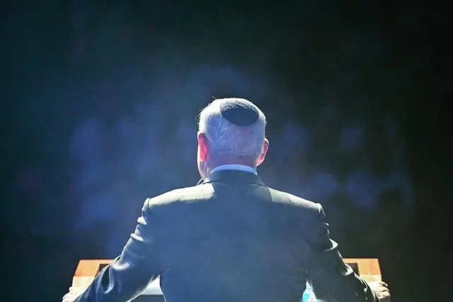 Spatele unui bărbat cu părul cărunt, purtând un costum închis la culoare și o kippa, fiind surprins în poziție de stat în fața unui podium.