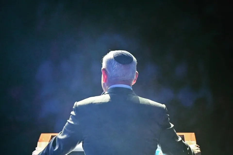 Spatele unui bărbat cu părul cărunt, purtând un costum închis la culoare și o kippa, fiind surprins în poziție de stat în fața unui podium.