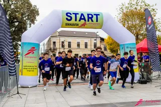 Mulți alergători în echipamente sportive trec pe sub o arcadă gonflabilă albă pe care scrie START.