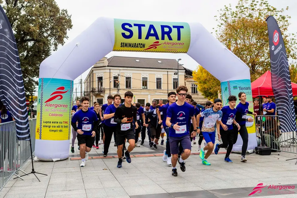 Mulți alergători în echipamente sportive trec pe sub o arcadă gonflabilă albă pe care scrie START.