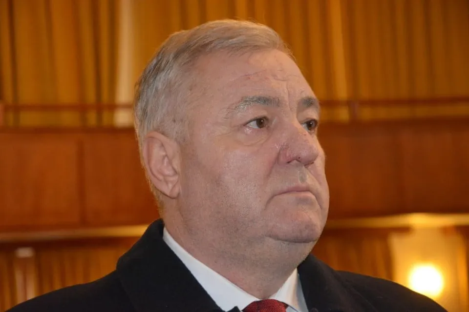 Un bărbat cu părul scurt și deschis la culoare poartă un sacou închis, o cămașă albă și o cravată roșie.