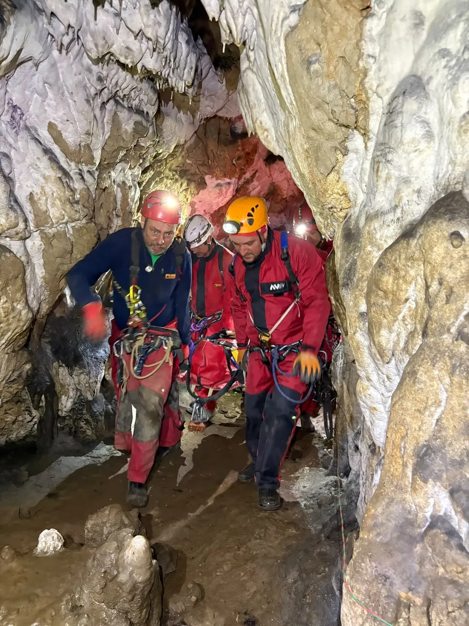 Trei persoane purtând căști de protecție și echipament de alpinism se deplasează printr-o peșteră cu pereți stâncoși și nisipoși.