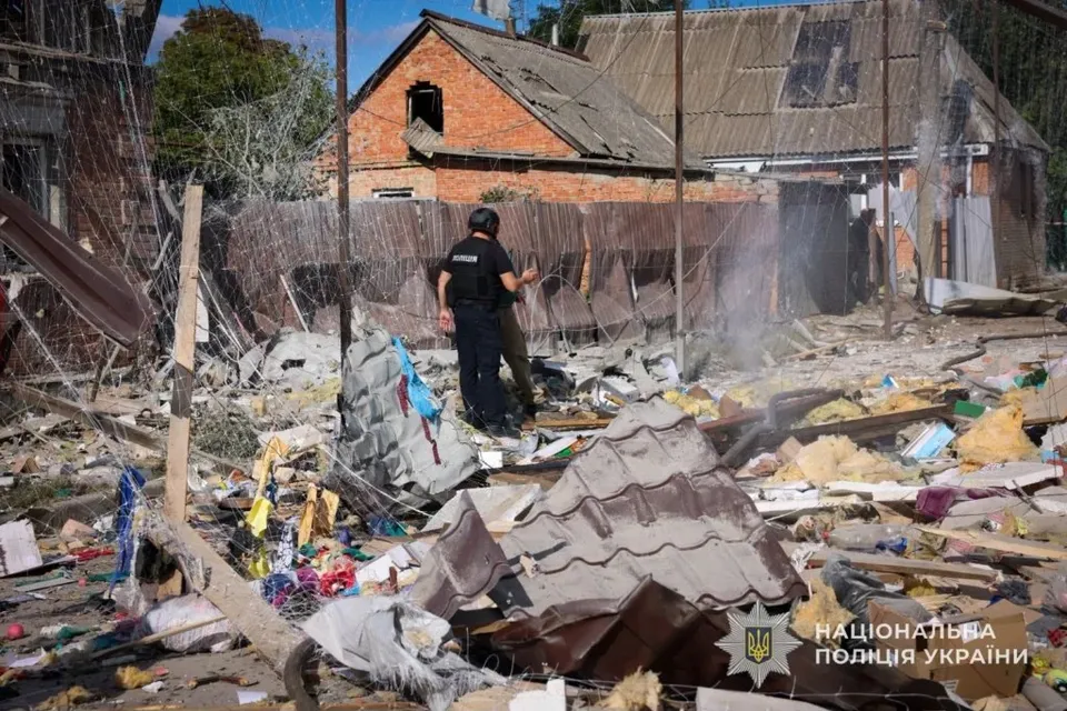 Un bărbat cu căști și haine închise stă în fața unor resturi de materiale și structuri de clădiri distruse sub un cer senin.