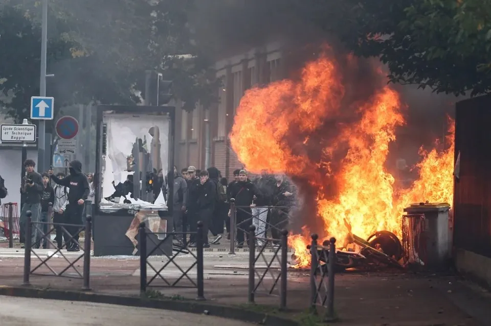 Flăcări mari de foc ard pe un trotuar lângă un coș de gunoi, în timp ce un grup de persoane stă în apropiere.