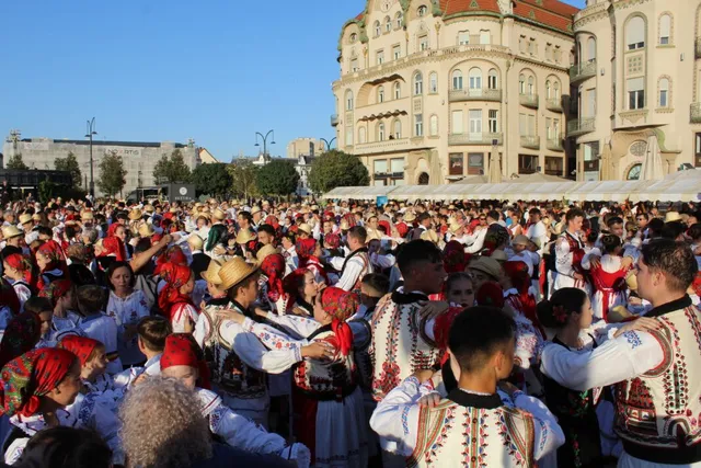 O mulțime de oameni îmbrăcați în costume populare dansează într-o piață publică cu clădiri înalte în fundal sub un cer senin.