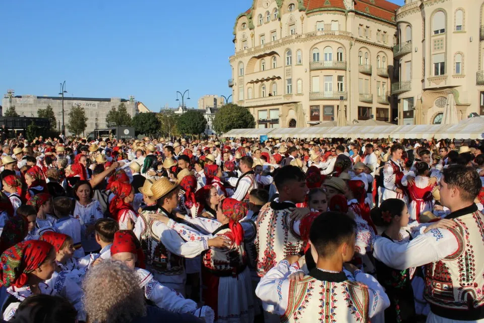 O mulțime de oameni îmbrăcați în costume populare dansează într-o piață publică cu clădiri înalte în fundal sub un cer senin.