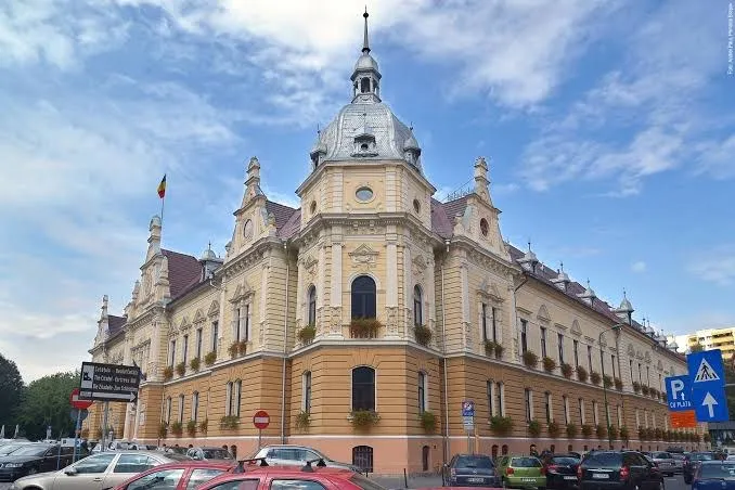 O clădire monumentală de culoare galben deschis cu acoperiș gri și turn central se află sub un cer cu nori, lângă un parc de mașini.