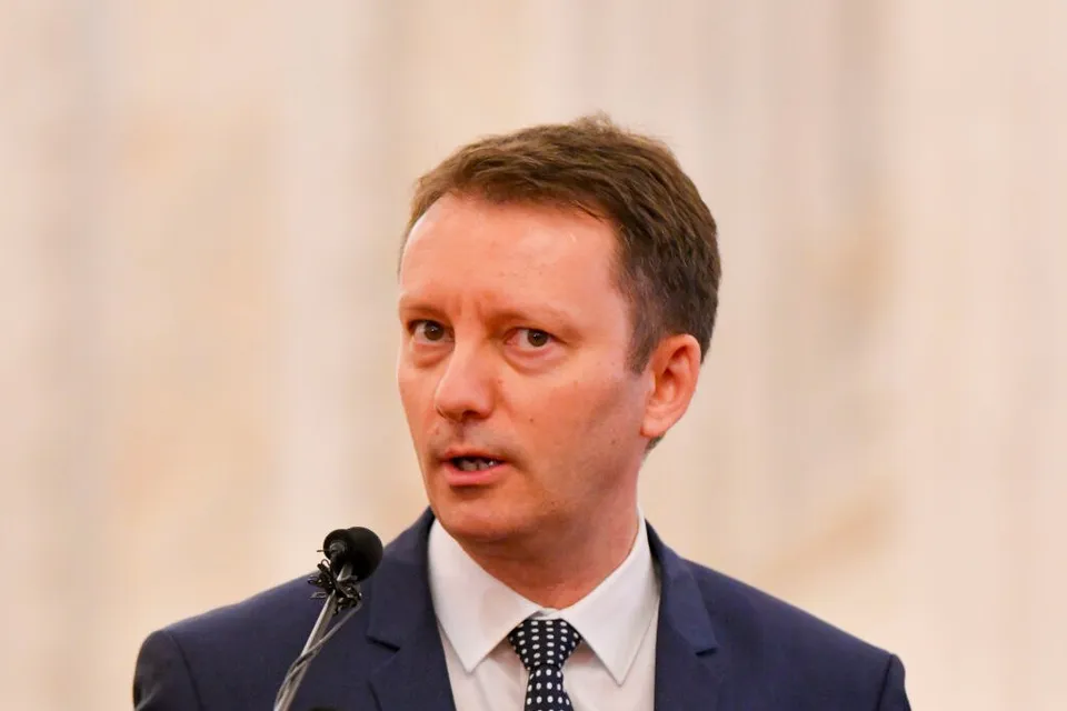 Un bărbat cu părul scurt și deschis la culoare poartă sacou albastru, cămașă albă și cravată cu puncte, vorbind în fața unui microfon.
