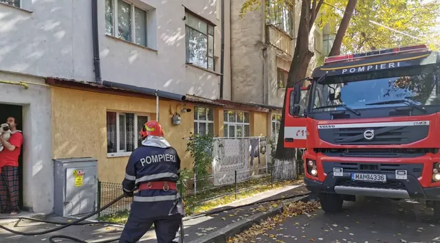 Un pompier cu echipament de protecție și un camion roșu sunt poziționate în fața unei clădiri de locuințe lângă un copac.