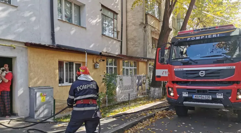 Un pompier cu echipament de protecție și un camion roșu sunt poziționate în fața unei clădiri de locuințe lângă un copac.