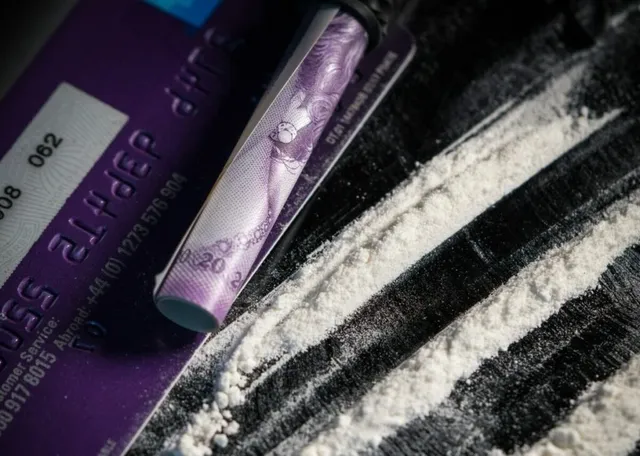Un tub de carton cu un desen violet este așezat lângă linii de o substanță albă granulară pe o suprafață închisă lângă un card violet.