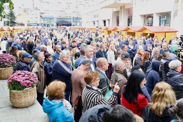 O mulțime de oameni stă într-o piață deschisă lângă rânduri de tarabe mici cu acoperișuri galbene și coșuri cu flori roz.