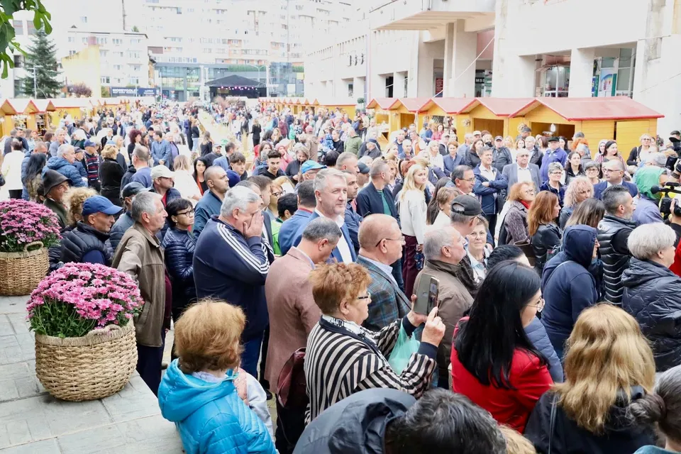 O mulțime de oameni stă într-o piață deschisă lângă rânduri de tarabe mici cu acoperișuri galbene și coșuri cu flori roz.
