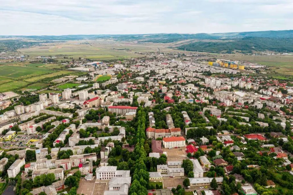 O vedere aeriană a unui oraș cu numeroase clădiri rezidențiale, zone verzi și terenuri deschise sub un cer înnorat.