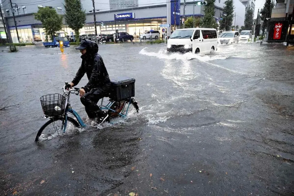 O persoană îmbrăcată în negru pedalează printr-o stradă inundată pe o bicicletă cu coș, în timp ce mașini circulă prin apă în fundal.