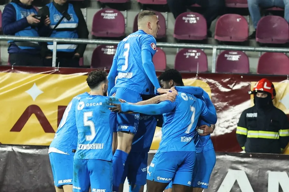 Câțiva jucători de fotbal în echipament albastru se îmbrățișează pe un teren de sport în fața unor tribune cu scaune roșii.