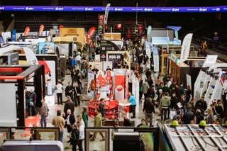 O mulțime de oameni circulă printre standuri de expoziție și structuri de prezentare într-o sală de sport cu tribune în fundal.