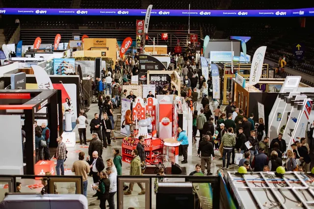 O mulțime de oameni circulă printre standuri de expoziție și structuri de prezentare într-o sală de sport cu tribune în fundal.