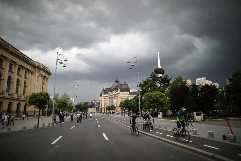 Câțiva bicicliști circulă pe o stradă asfaltată sub un cer acoperit de nori întunecați, lângă clădiri și copaci.