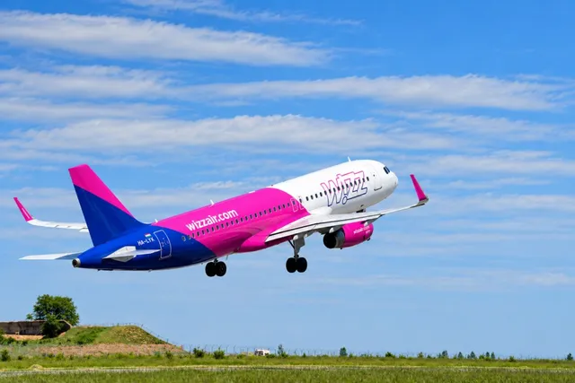 Un avion de pasageri roz și alb zboară deasupra unui câmp verde sub un cer albastru cu nori albi.