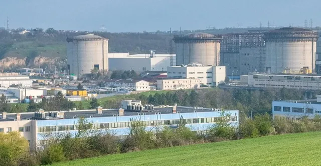 Mai multe clădiri industriale cilindrice și rectangulare sunt situate într-un peisaj cu vegetație și dealuri sub un cer senin.