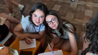 Două tinere stau la o masă de lemn, purtând haine de vară, și scriu pe foi de hârtie cu pixuri.