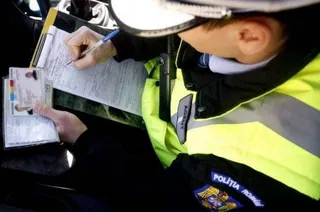 Un agent de poliție în uniformă portivită cu benzi reflectorizante scrie pe o foaie de hârtie de pe un dosar, ținând un permis de conducere.