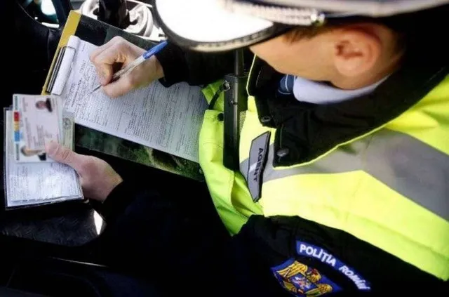 Un agent de poliție în uniformă portivită cu benzi reflectorizante scrie pe o foaie de hârtie de pe un dosar, ținând un permis de conducere.