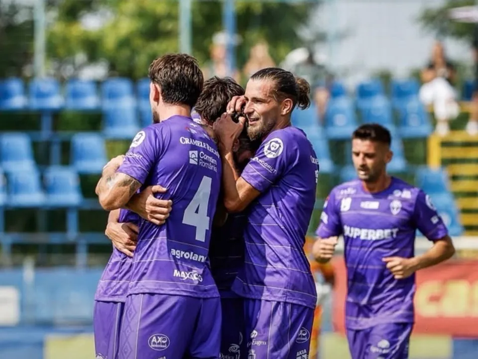 Patru jucători de fotbal îmbrăcați în echipamente mov se îmbrățișează pe un teren de sport sub lumina soarelui.