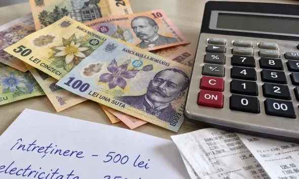 Un calculator gri stă lângă bancnote românești și câteva hârtii scrise cu text manuscris pe o suprafață plană.