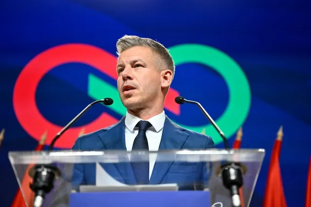 Un bărbat cu părul scurt și deschis la culoare, purtând sacou albastru și cravată închisă, stă în spatele unui podium cu microfoane.