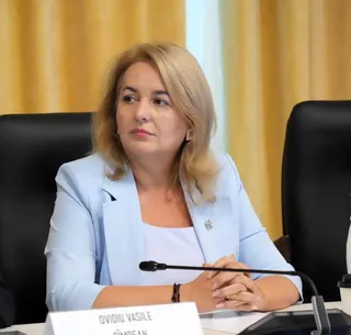 O femeie cu părul blond, purtând un sacou bleu și un tricou alb, stă la o masă cu mâinile încrucișate.