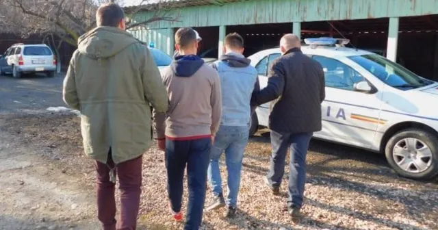 Patru bărbați merg pe un drum de pietriș lângă o mașină de poliție albă și o clădire cu acoperiș verde.