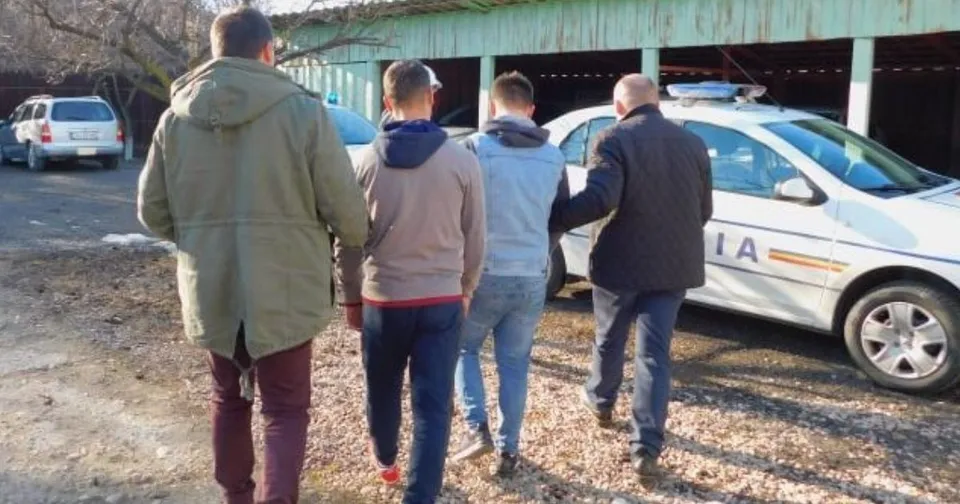 Patru bărbați merg pe un drum de pietriș lângă o mașină de poliție albă și o clădire cu acoperiș verde.