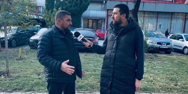 Un bărbat cu barbă și un bărbat cu microfon stau pe un covor de iarbă lângă mașini parcate.