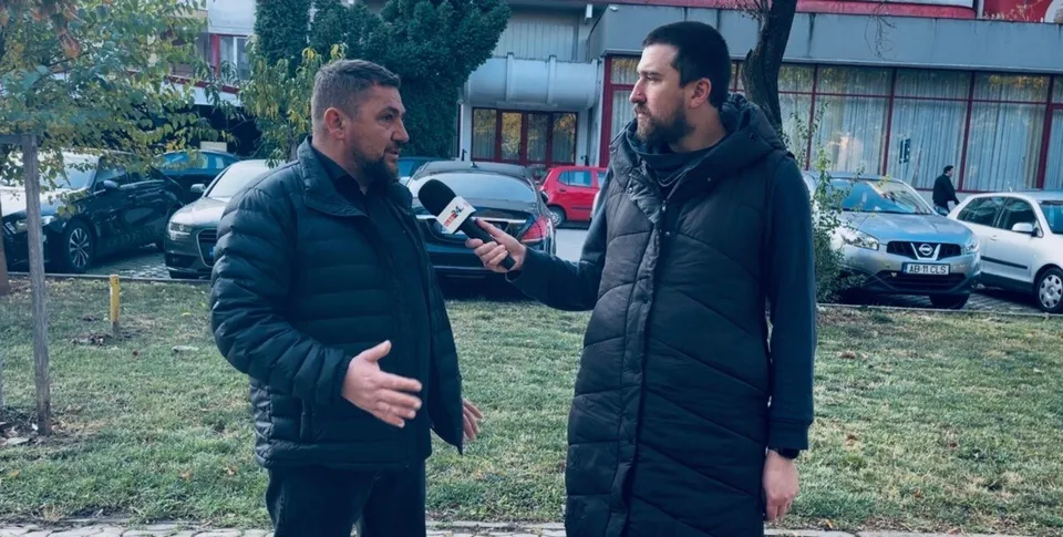 Un bărbat cu barbă și un bărbat cu microfon stau pe un covor de iarbă lângă mașini parcate.