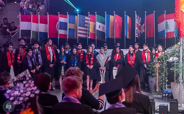 Un grup de absolvenți în robe și toci, cu eșarfe roșii și albastre, stau în fața unui rând de steaguri internaționale, în timpul unei ceremonii.