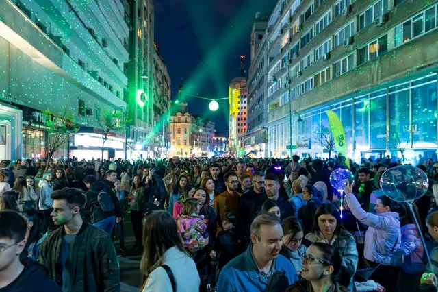 O mulțime de oameni stă pe o stradă urbană noaptea, sub proiecții de lumină verzi și albastre pe clădirile din fundal.