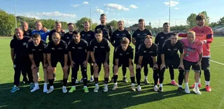 Un grup de fotbaliști în echipamente sportive negre și roz pozează într-o poziție de grup pe un teren de fotbal verde sub cerul senin.