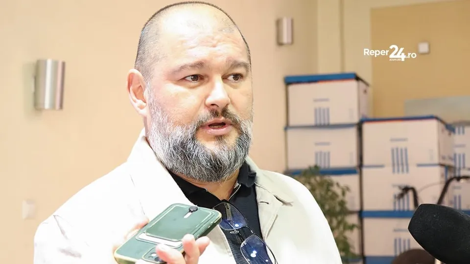 Un bărbat cu barbă și păr scurt, purtând o jachetă deschisă, ține un telefon mobil în mână în fața unor cutii albe stivuite.