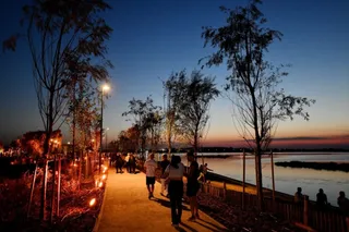 Oamenii merg pe un drum pietonal lângă un lac, sub siluete de copaci sub un cer de seară.