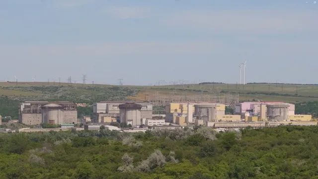 O serie de clădiri industriale și structuri de beton sunt situate într-un peisaj cu vegetație verde sub un cer senin.