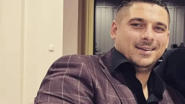 Un bărbat cu părul scurt și barbă stufoasă poartă un sacou cu model de carouri și o cămașă neagră, zâmbind spre cameră.