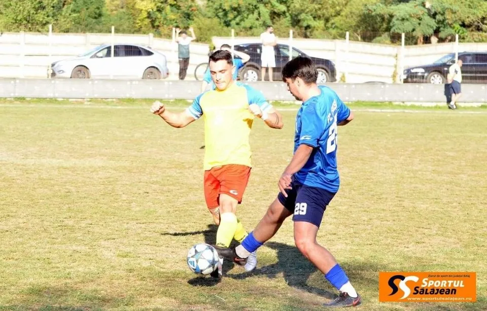 Un jucător în echipament albastru lovește un fotbal pe un teren de iarbă, în fața unui alt jucător îmbrăcat în galben și roșu.