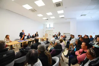 Un grup de persoane stă așezat pe scaune într-o încăpere cu pereți albi, privind spre un bărbat care stă în spatele unei mese lungi.