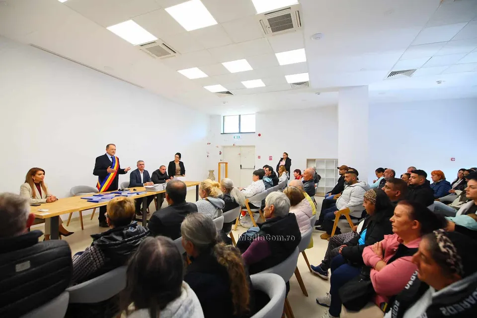 Un grup de persoane stă așezat pe scaune într-o încăpere cu pereți albi, privind spre un bărbat care stă în spatele unei mese lungi.