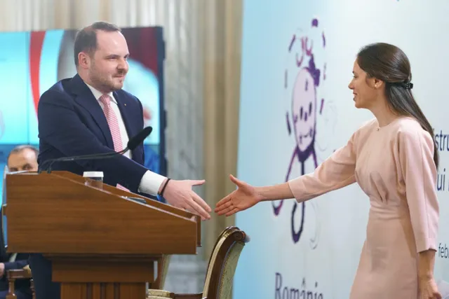 Un bărbat într-un costum închis și o femeie într-o rochie roz se întind spre o strângere de mână în fața unui podium de lemn.