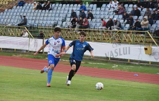 Doi jucători de fotbal de pe un teren de gazon verde aleargă spre un balon alb.
